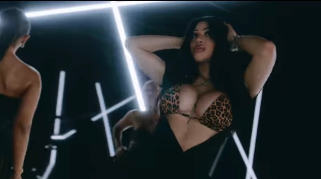 A woman wearing a leopard print bikini top poses with her hands behind her head, showcasing confidence and attitude against a backdrop of neon lights.